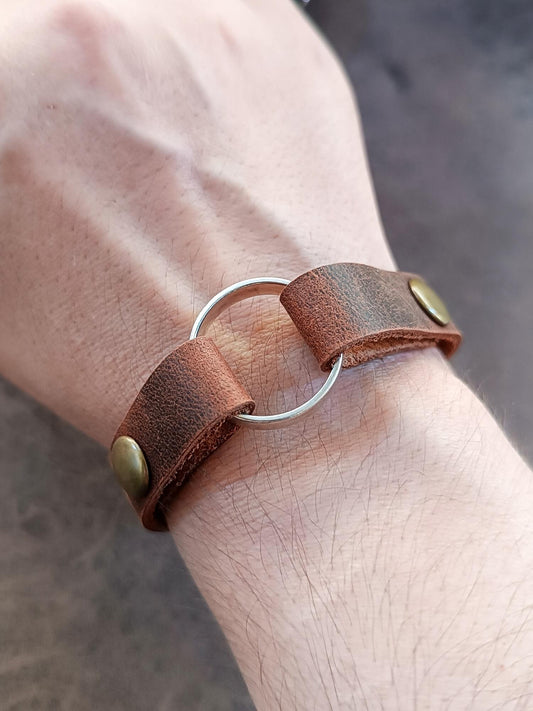 Minimalist leather ring holder cuff (1.2 cm) in Oak Brown worn on a wrist, securely holding a silver wedding band. Focus on slim profile.