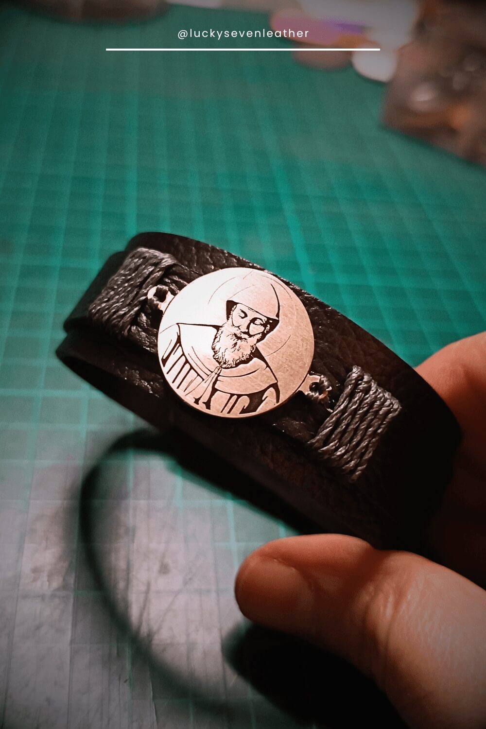 Leather bracelet with a circular emblem featuring St. Charbel, held by a hand on a green cutting mat.