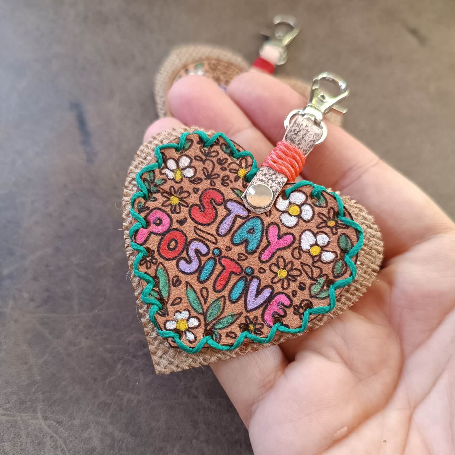 Detailed close-up of the hand-stitched "Stay Positive" Leather Heart Keychain, highlighting the vibrant hand-painted floral design, colorful bubble font, and intricate green waxed cord stitching.