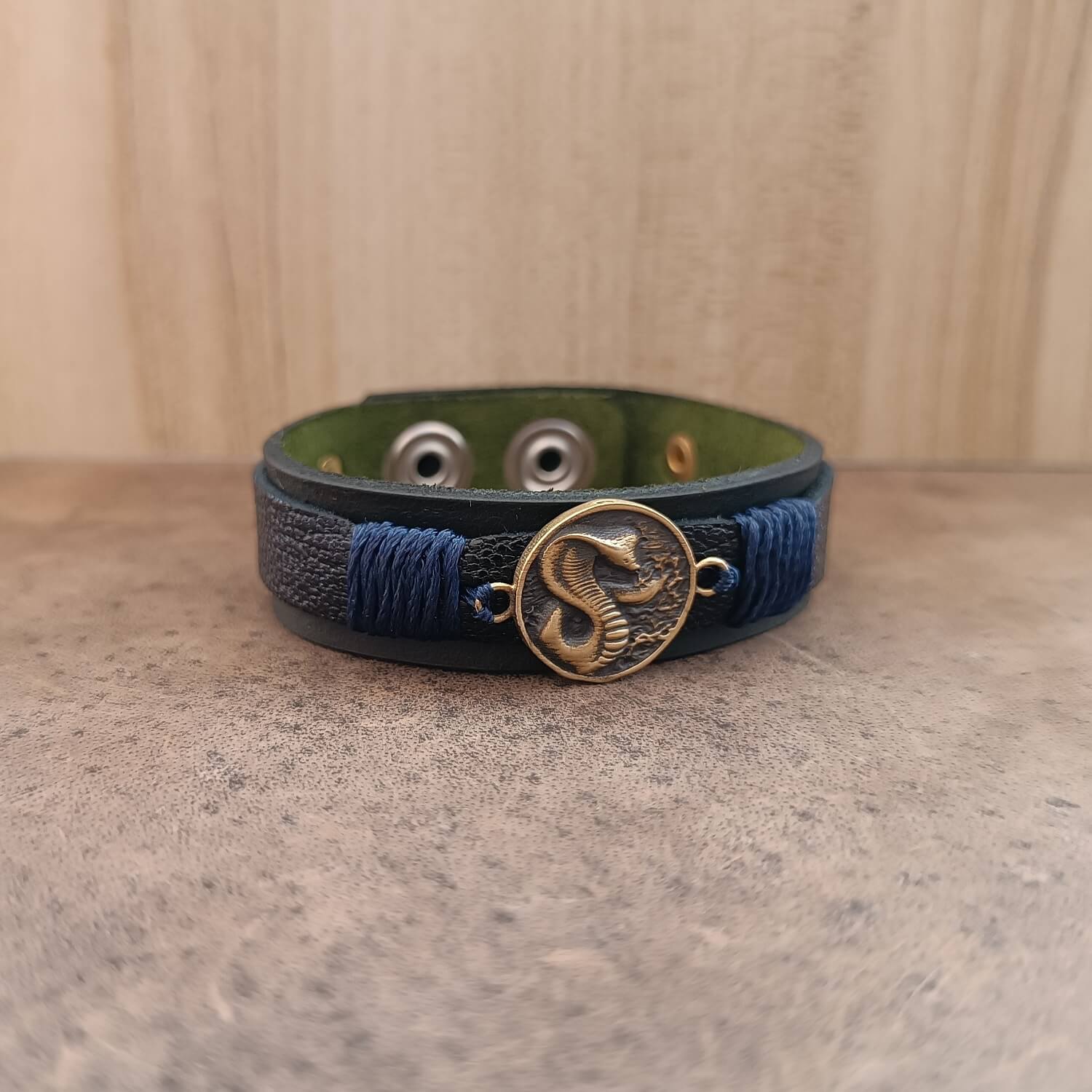 Close-up front view of the Vincenzo Bracelet Cobra Edition showing the matte black full-grain leather base and crackled black top strip. Centered oxidized brass cobra pendant gleams with subtle golden shadows, framed by seven navy blue cord wraps on each side, symbolizing silent strength and strategic patience.