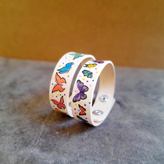 Two white leather bracelets with colorful hand-painted butterfly patterns and polka dots, displayed on a textured background.