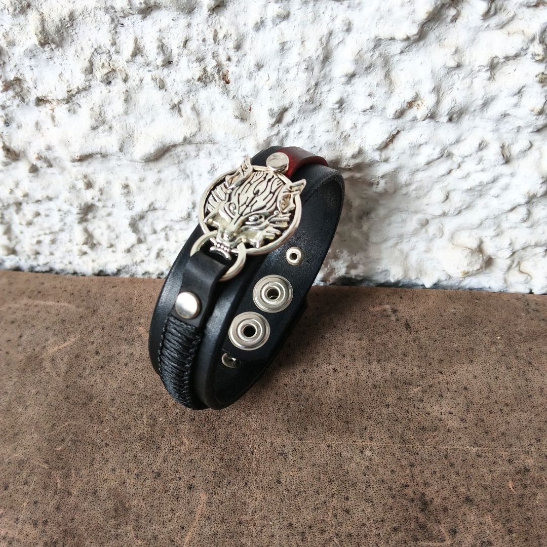 A black leather wrap bracelet with a silver-toned metal tree of life charm, rivets, and a dark red inner lining, displayed on a brown textured surface against a white stucco wall.