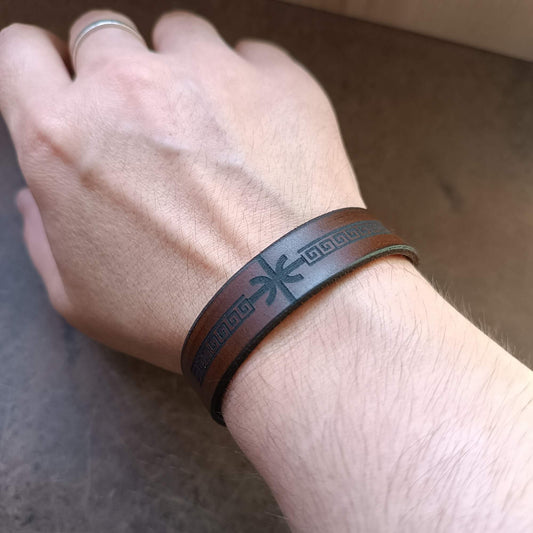 Hand wearing a brown leather bracelet with delphic epsilon and meander design on a wooden surface