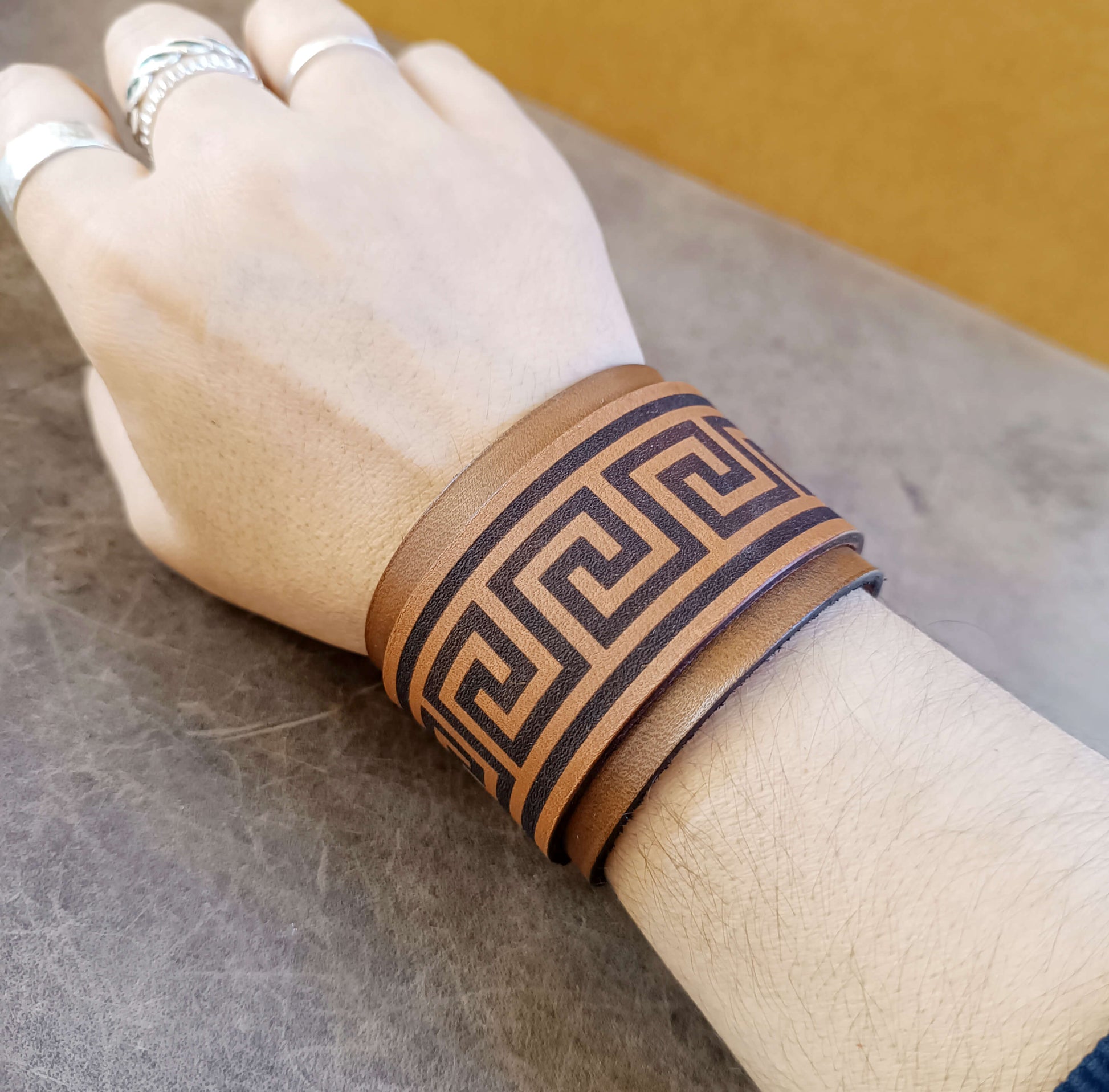 Hand wearing a brown leather cuff with Greek key design on a neutral background