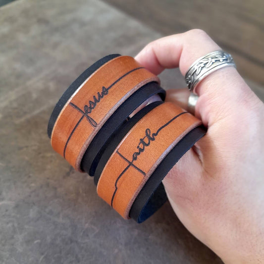 The Freedom Leather Cuff Bracelet with "Faith" deeply engraved in calligraphy and "Jesus" variation held on hand.
