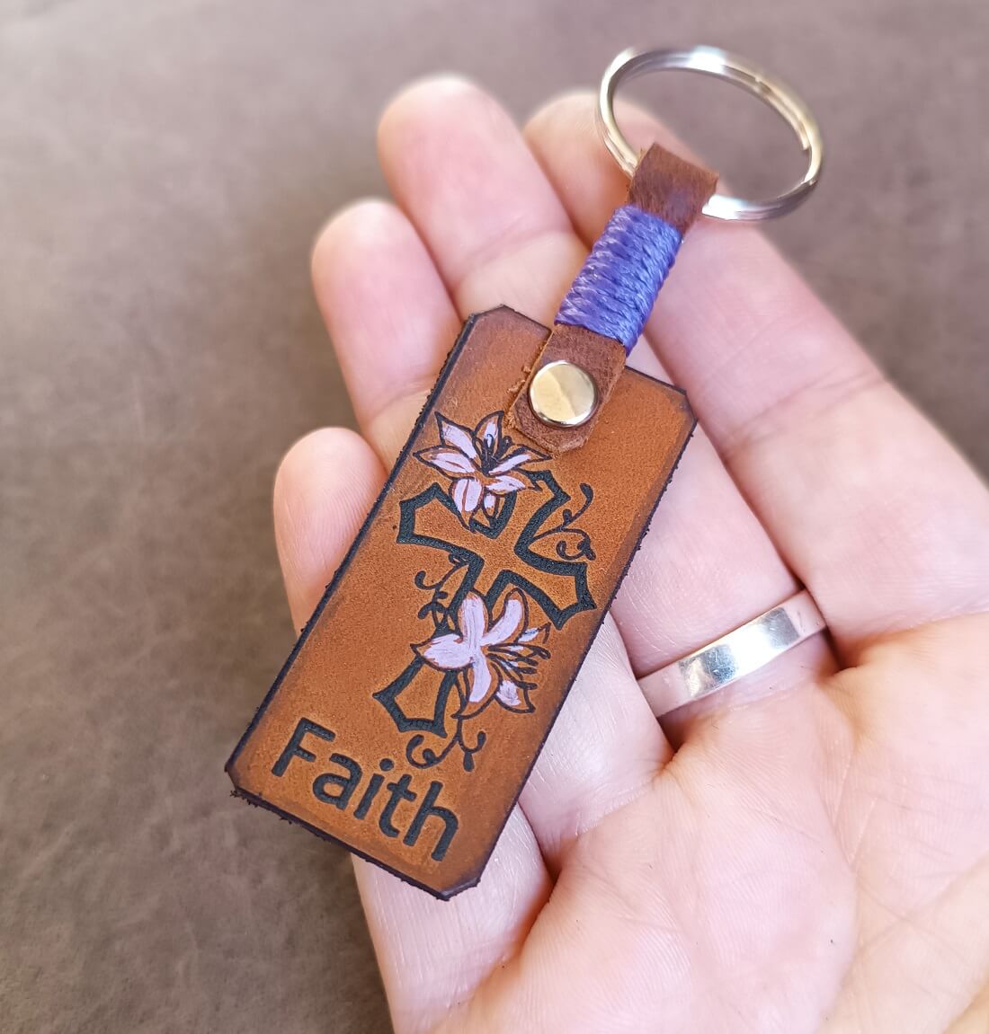 A brown leather keychain featuring a laser engraved and hand-painted floral cross with the word 'Faith' below it. The keychain also includes a personalized element, which appears to be a name in cursive script.