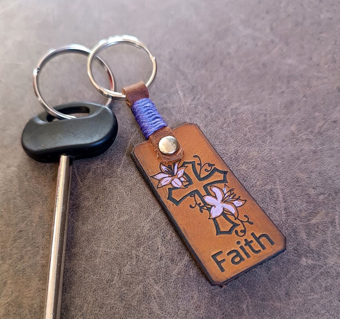 A brown leather keychain featuring a laser engraved and hand-painted floral cross with the word 'Faith' below it. The keychain also includes a personalized element, which appears to be a name in cursive script.