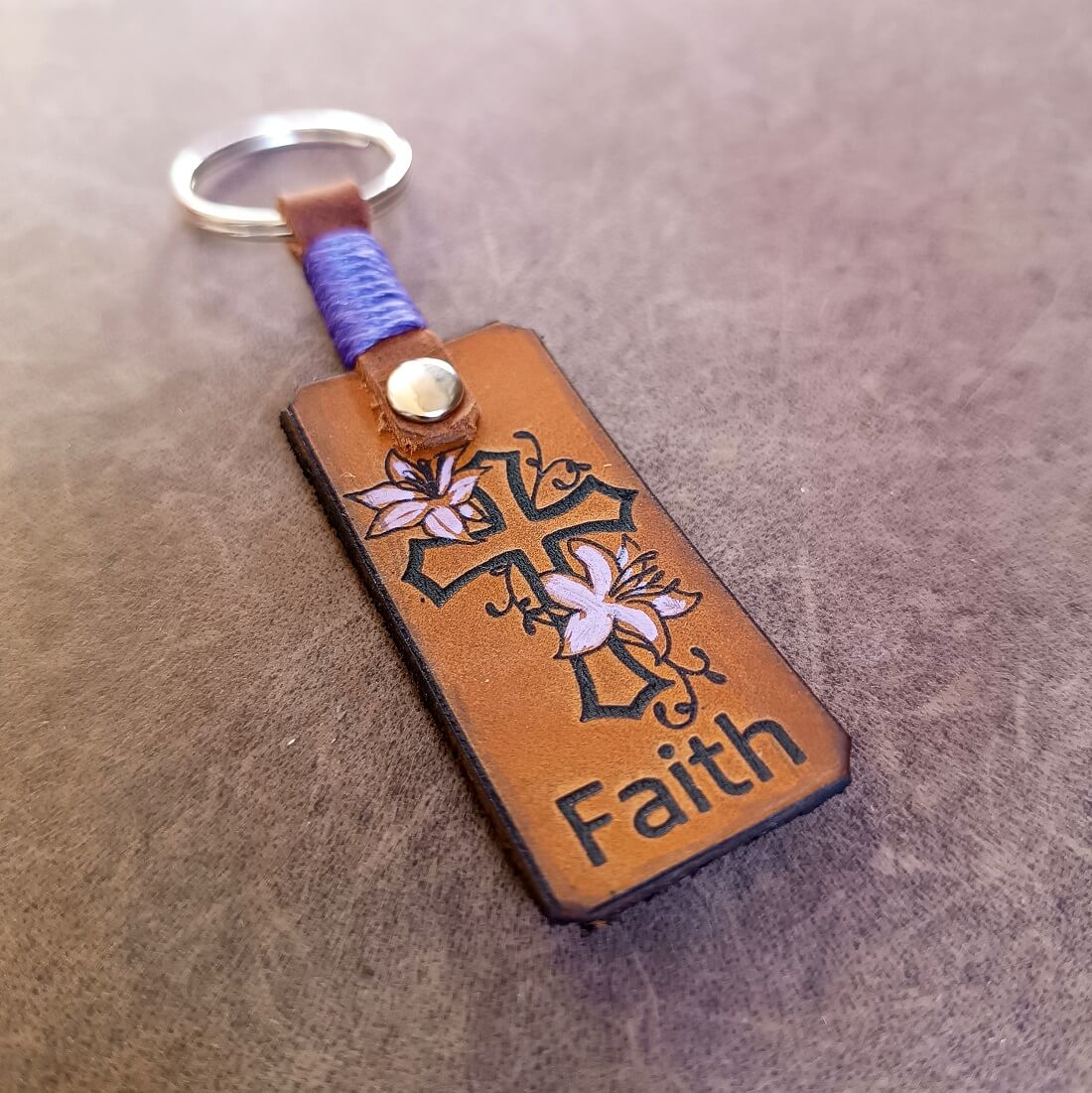 A brown leather keychain featuring a laser engraved and hand-painted floral cross with the word 'Faith' below it. The keychain also includes a personalized element, which appears to be a name in cursive script.