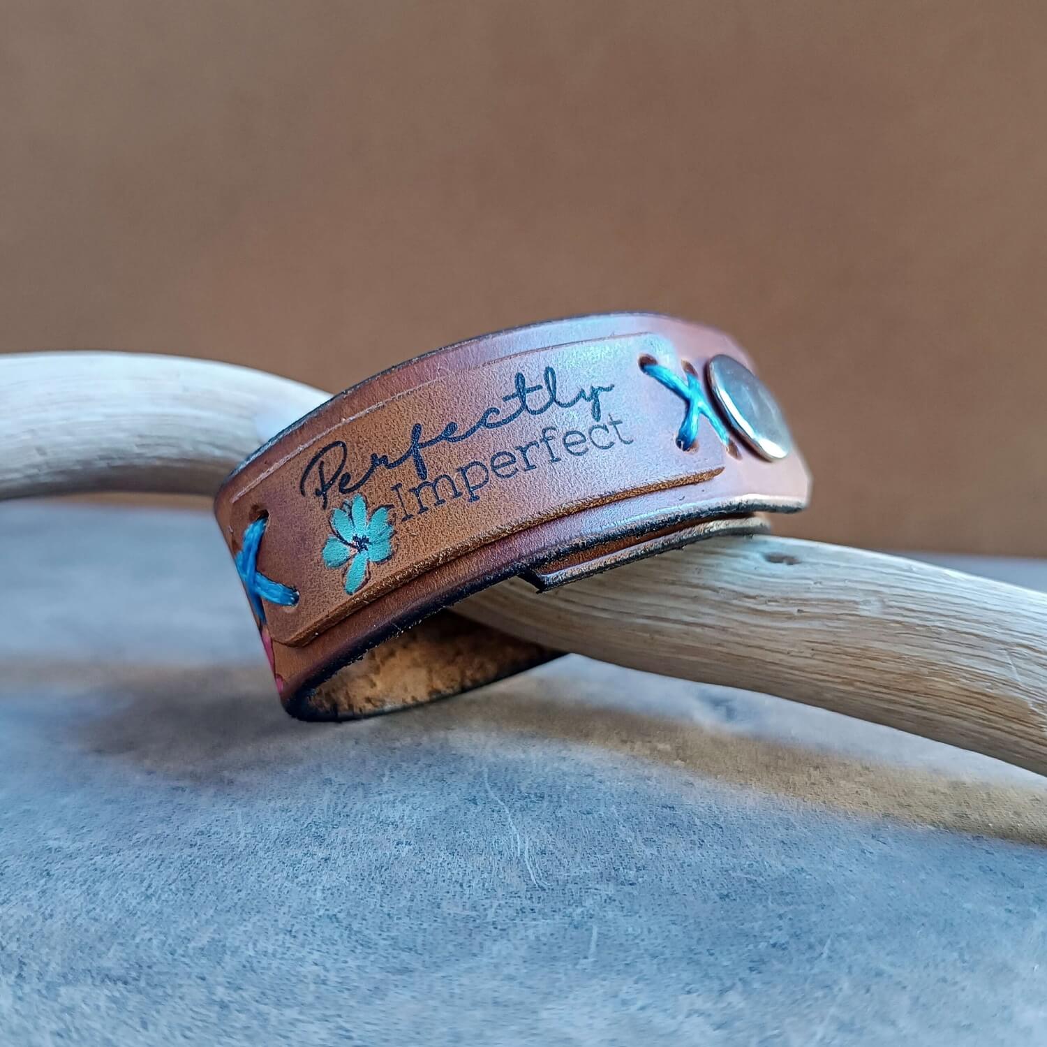 The Floral lioness Brown leather bracelet with 'Perfectly Imperfect' text close to the clasp and blue thread on a wooden surface.