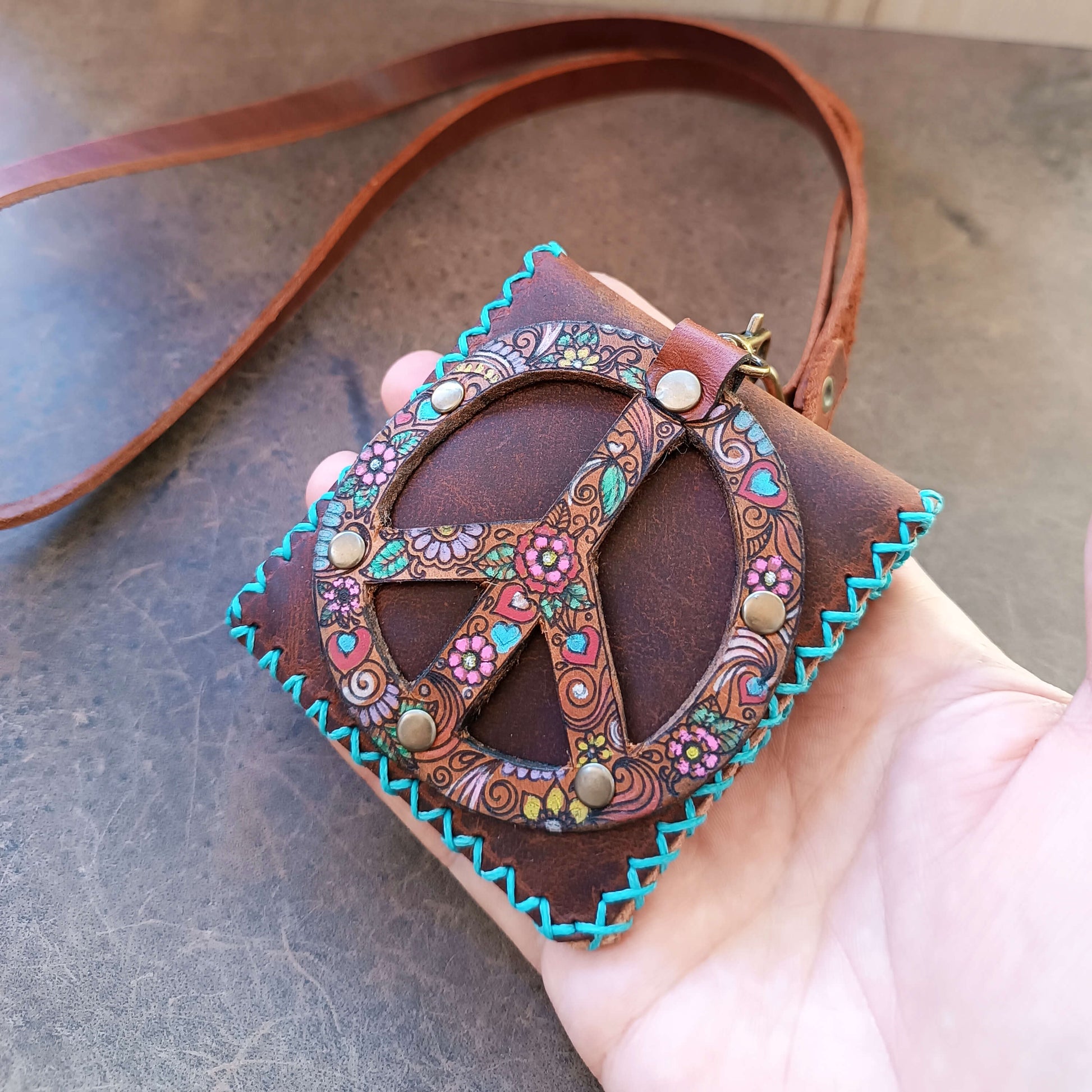 Decorative leather neck bag with peace symbol design held by a hand on a textured surface
