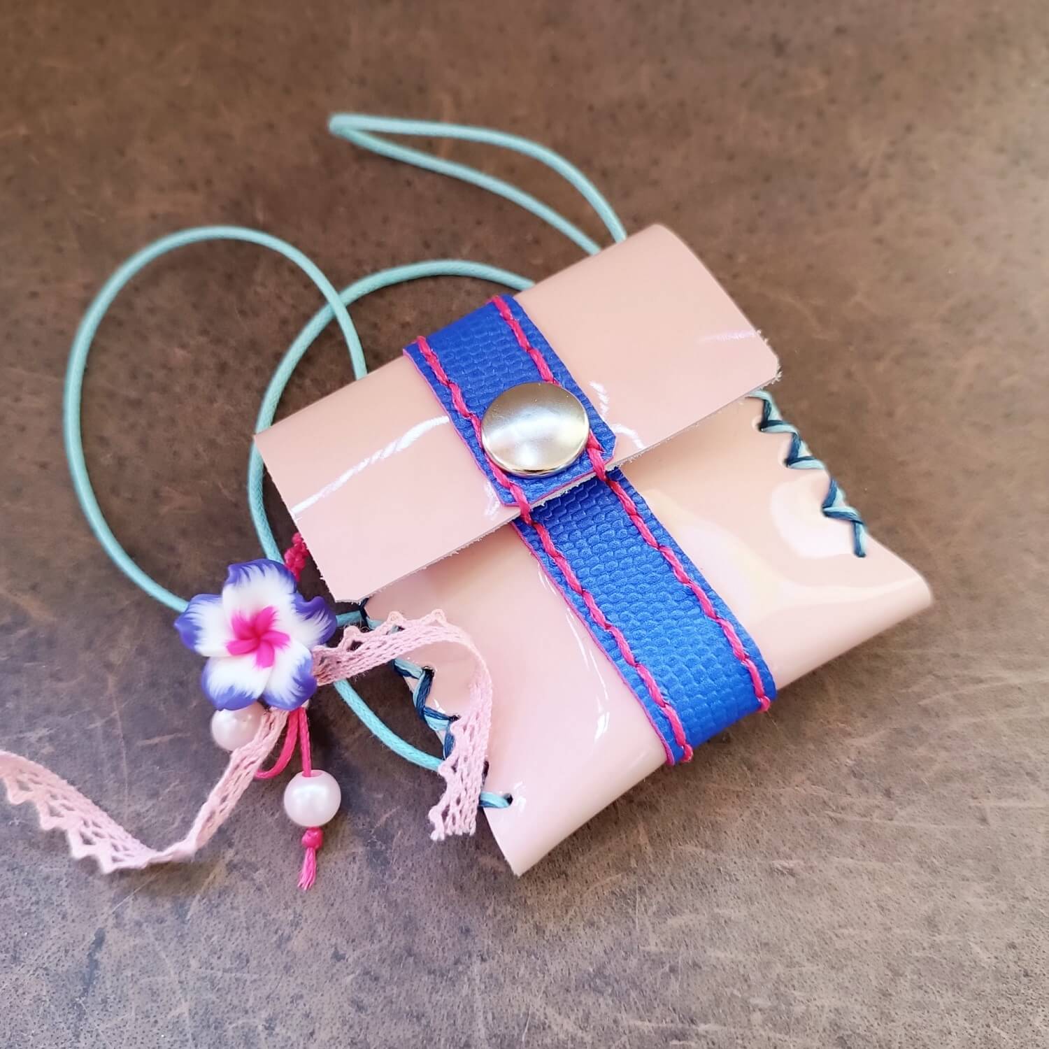 A glossy pink patent leather necklace coin purse with a blue/purple ombre leather strap, a polymer clay flower charm, and a light blue cotton cord.