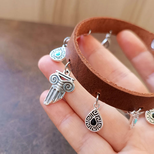 Hand holding a brown leather anklet with various silver greek charms.