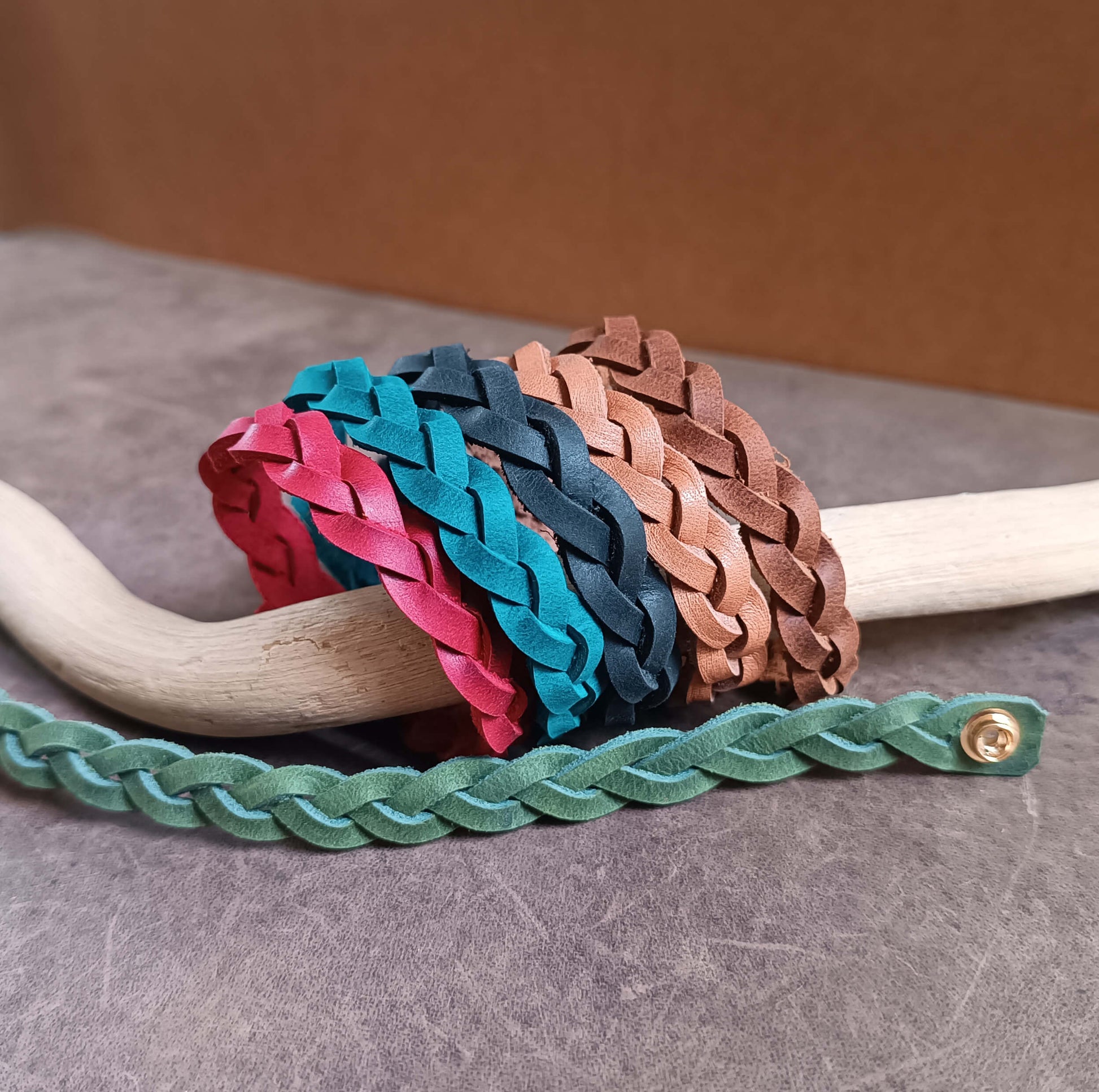 Six braided leather bracelets in red, blue, brown, chocolate, black and green on a wooden stick with a gray background.