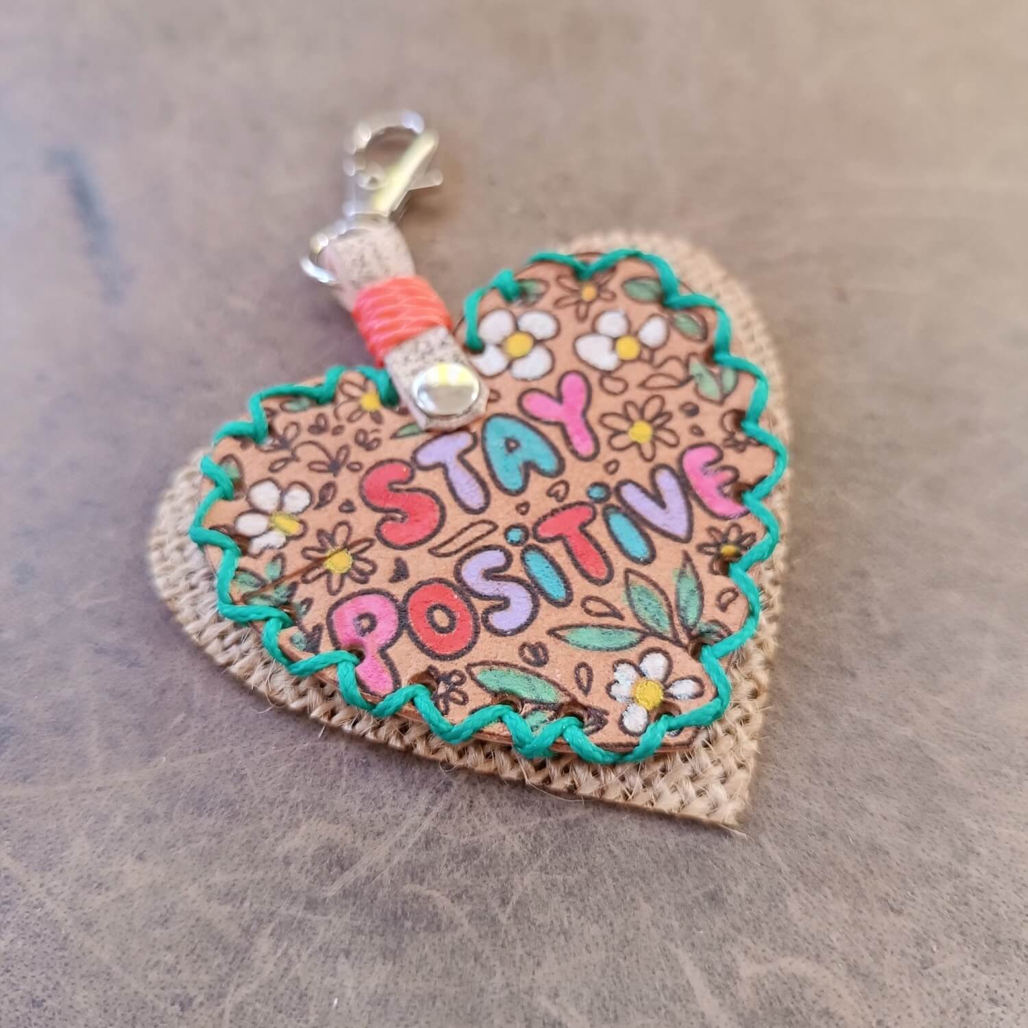side view of the hand-stitched "Stay Positive" Leather Heart Keychain, highlighting the vibrant hand-painted floral design, colorful bubble font, and intricate green waxed cord stitching.