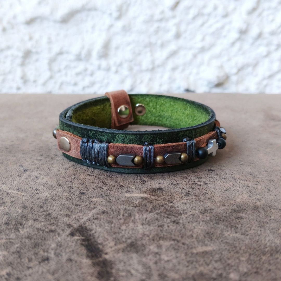 A handmade leather wrap bracelet with a distressed green and brown finish, featuring brass studs, a dark gray thread wrap, and small arrow accents, resting on a textured brown surface.
