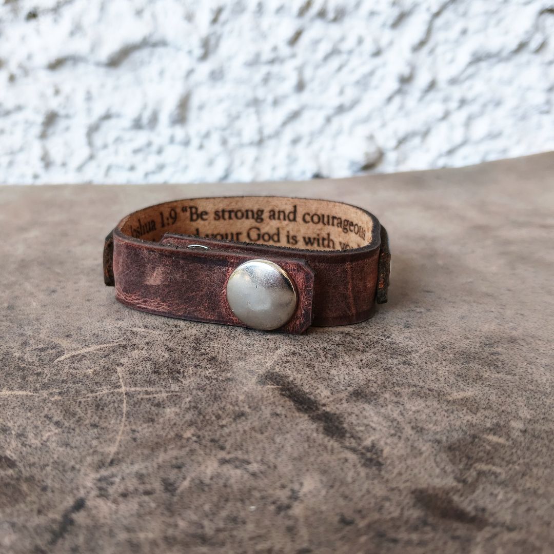 A distressed brown leather cuff bracelet with a silver-toned metal snap closure and engraved Bible verse text inside, resting on a textured brown surface against a white stucco wall.