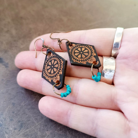 A pair of boho style dangle earrings with a geometric mandala design, featuring turquoise czech beads and antique bronze hooks, held in a person's hand.