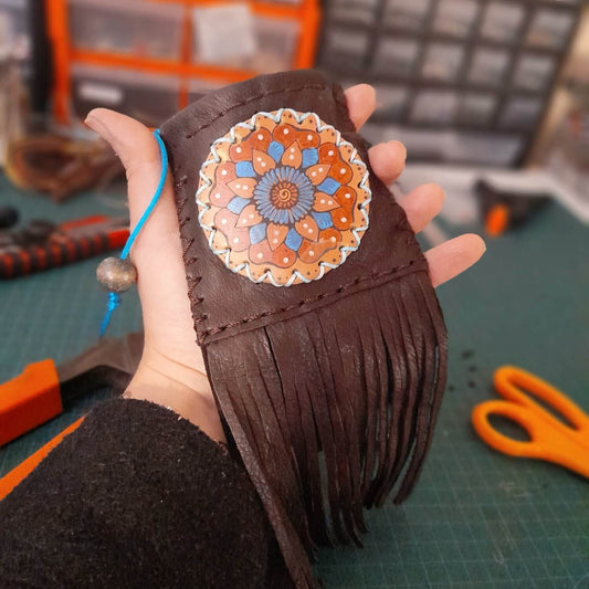 A small dark brown leather pouch with a hand-painted mandala design in blue and red, adorned with fringe and a drawstring closure.