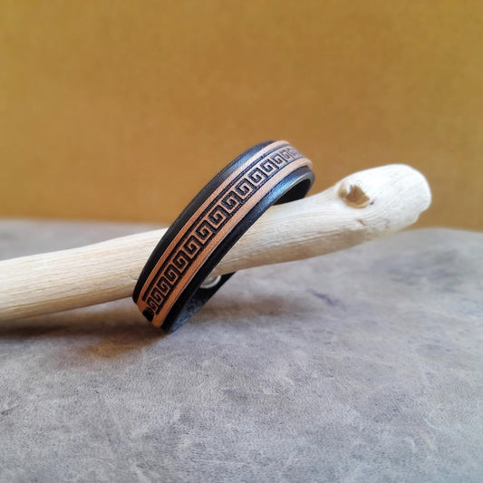 A black leather bracelet with a Greek Meander symbol engraved on it, displayed on a wooden surface.