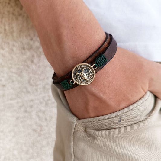 Close-up of a wrist wearing a brown leather and green cord bracelet with a medusa pendant on a neutral background.