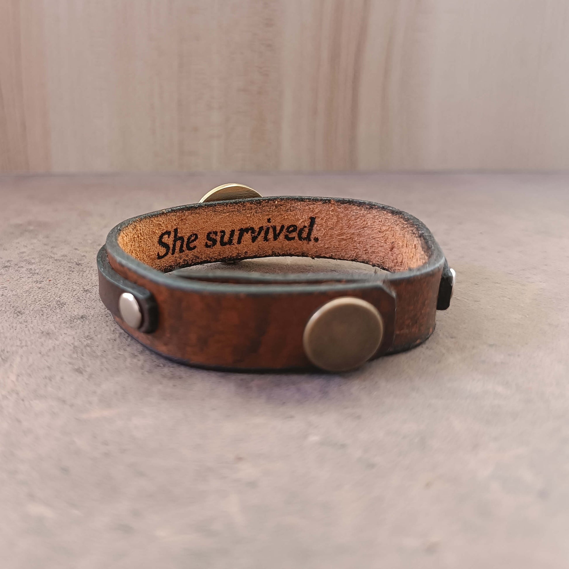 Brown leather bracelet with 'She survived.' engraving on a neutral background