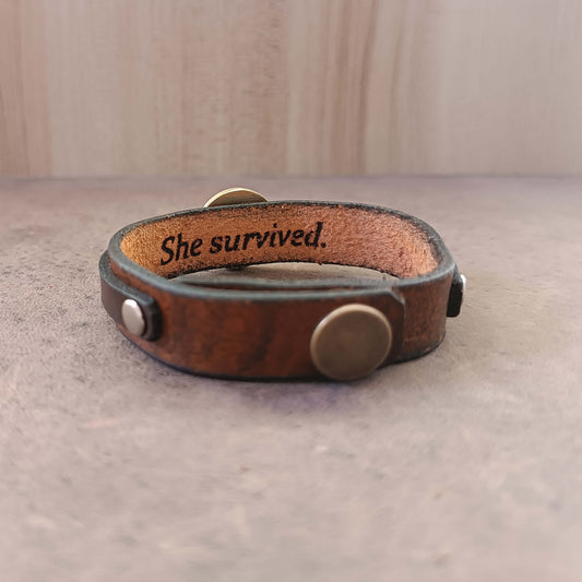 Brown leather bracelet with 'She survived.' engraving on a neutral background