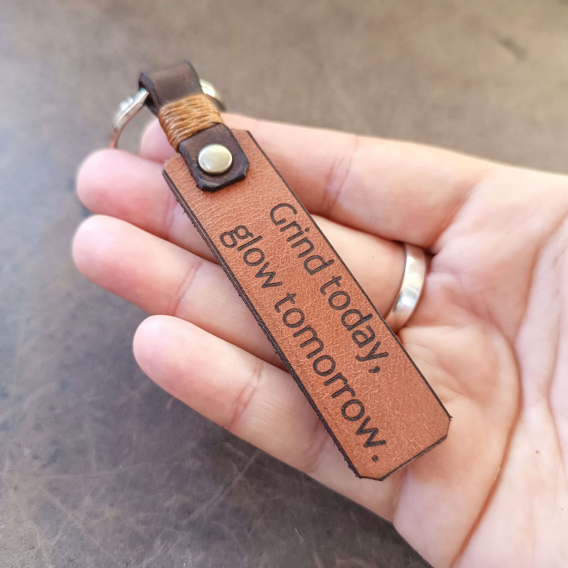 Brown leather keychain with 'Grind today, glow tomorrow' text held in a hand.