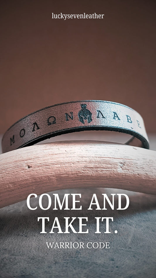 Leather bracelet with engraved text on a wooden surface, featuring the phrase 'COME AND TAKE IT.'