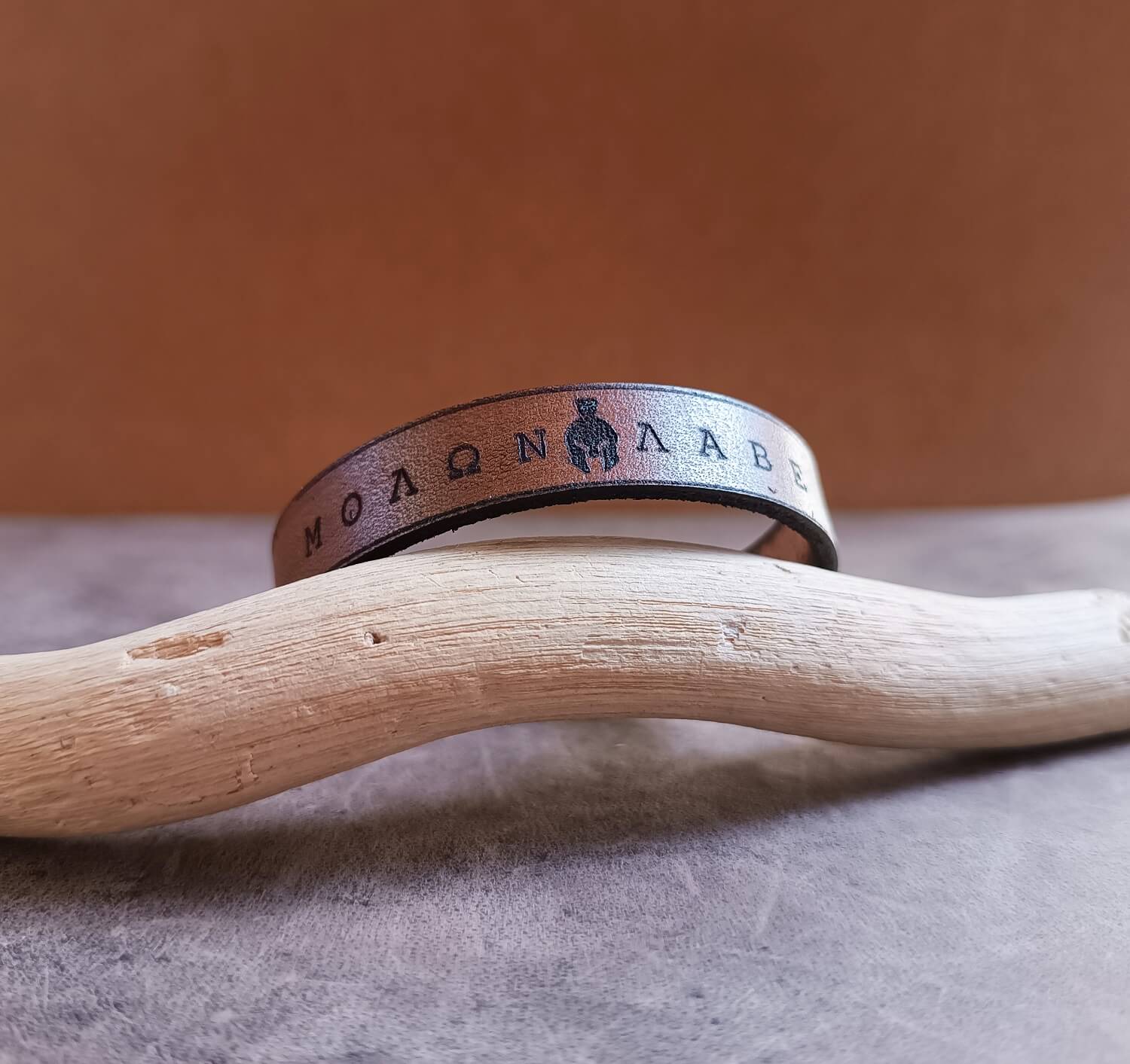 Molon Lave brown leather bracelet with engraved text on a wooden branch against a brown background