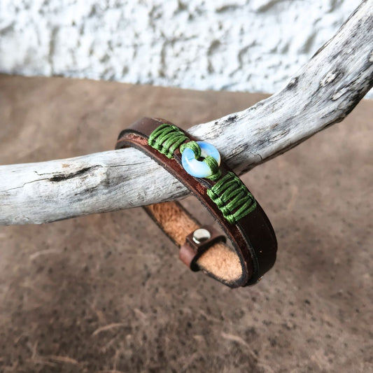 A handmade leather wrap bracelet with green braided cord, a moonstone donut bead, and a metal snap closure, resting on a weathered branch over a textured brown surface.