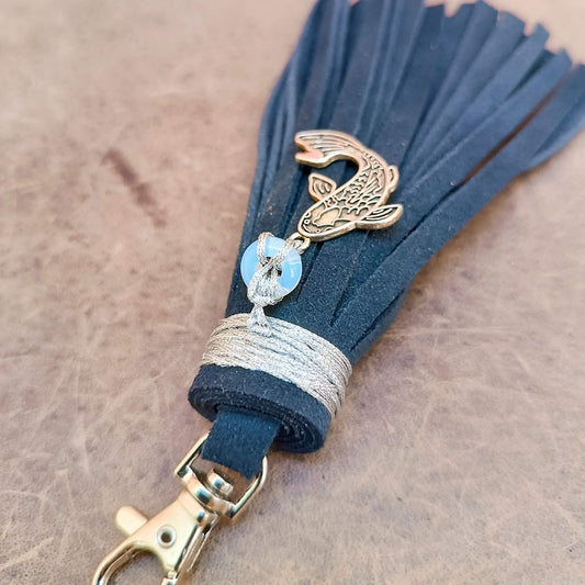 A dark blue suede tassel keychain featuring a koi fish charm, a lmoonstone donut bead with a white knot, and a silver wrapped cord, attached to a silver clasp on a textured brown surface.