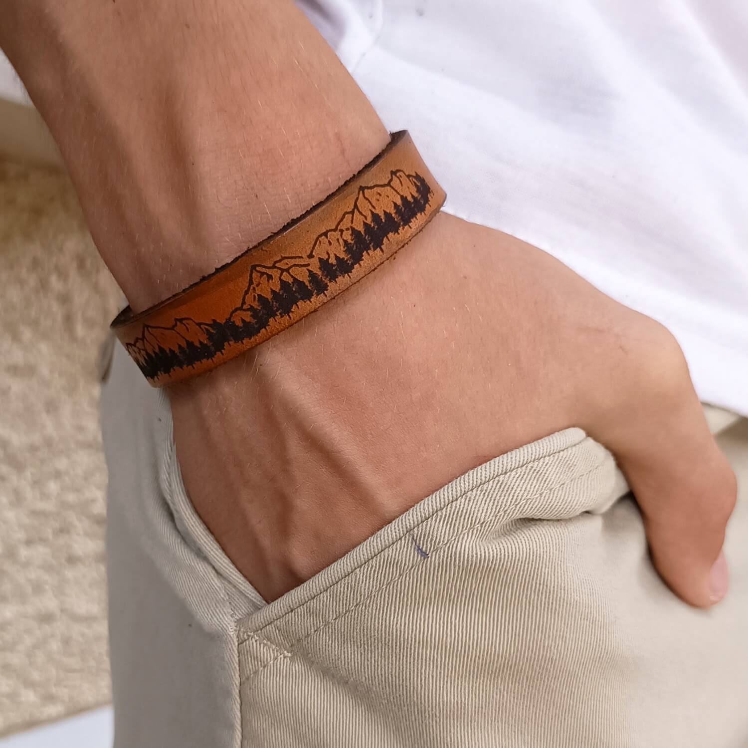 Brown leather bracelet with mountains and trees design on a wrist, worn by a person.