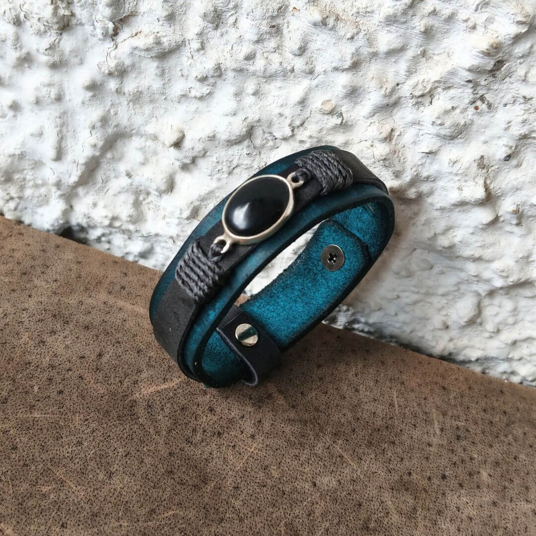A multi-layered wrap bracelet with a teal leather base, black braided cord, a black oval stone, and silver-toned metal accents, resting on a brown textured surface against a white stucco wall.