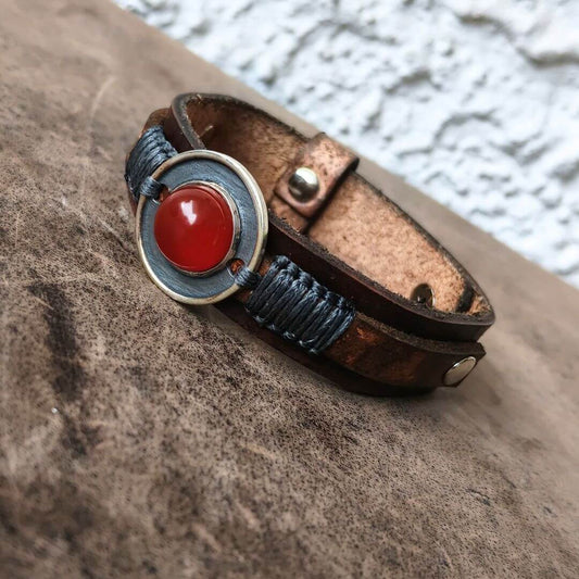 A handmade leather wrap bracelet with a central round bezel containing a red cabochon stone, accented by blue braided cord and brass hardware, resting on a textured brown surface.