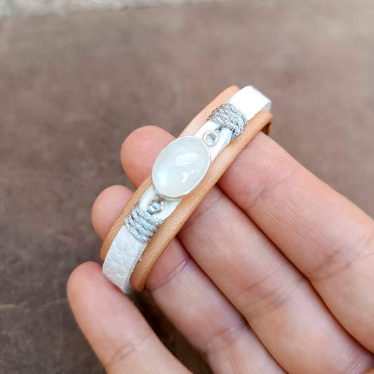 A close-up of a hand holding a double-strand leather bracelet with a white pebbled-leather band, a tan leather band, silver cord wrapping, and a central oval moonstone.