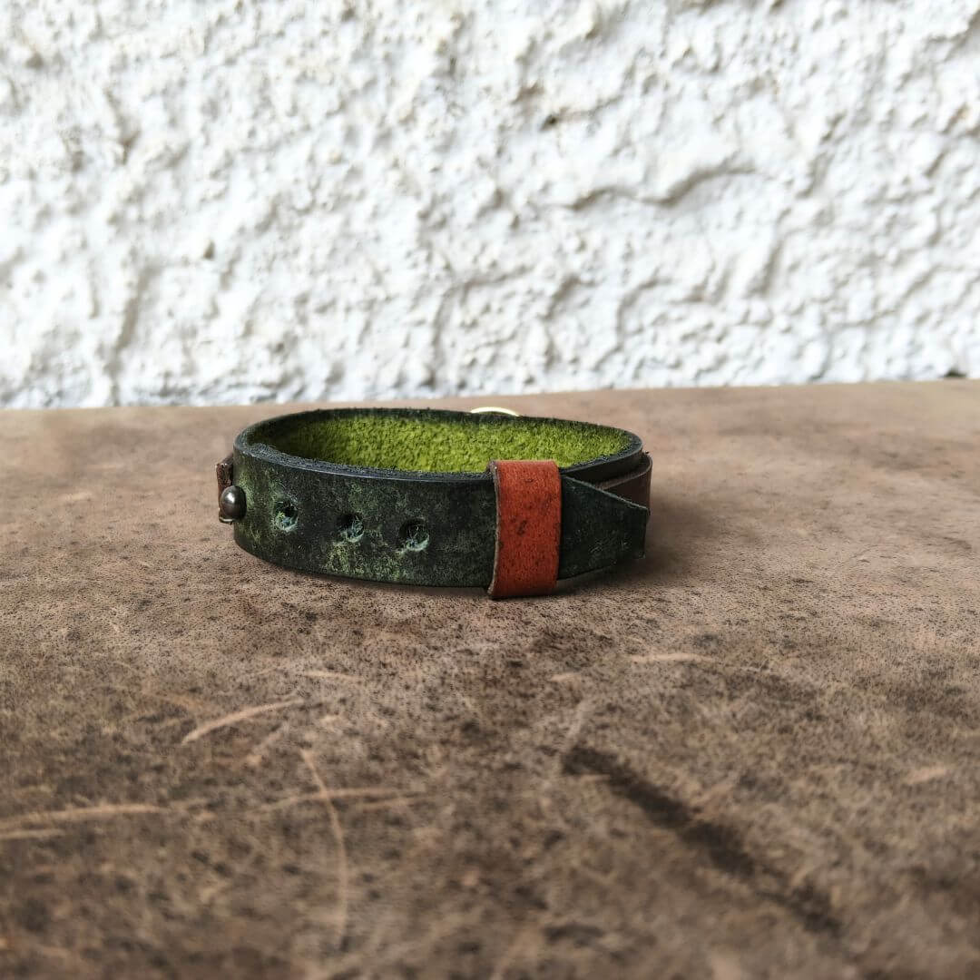 A handmade leather cuff bracelet with a distressed dark green finish, a bright green interior lining, and a contrasting reddish-brown leather tab, resting on a textured brown surface against a white stucco wall.