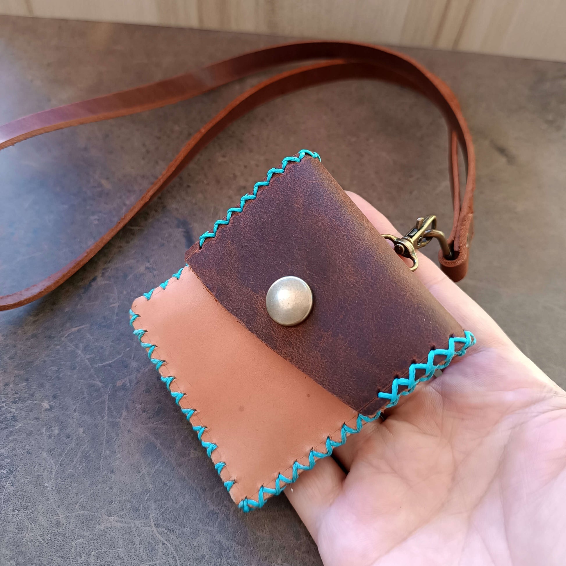 Handmade leather pouch with turquoise stitching held by a hand on a textured surface.