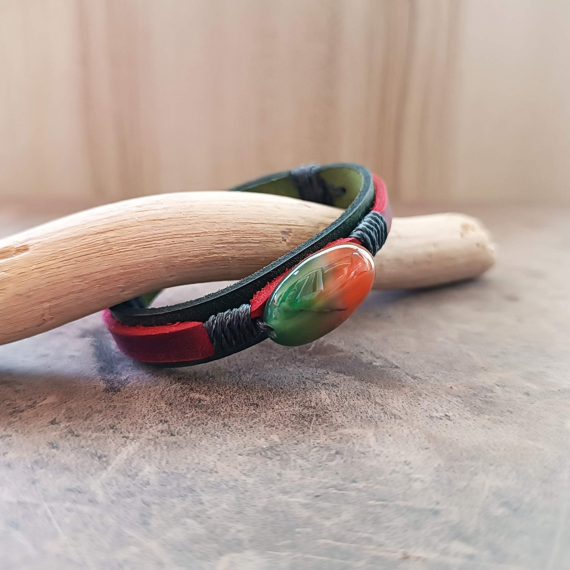 green and red leather Bracelet with a colorful peacock agate bead on a wooden stick against a neutral background
