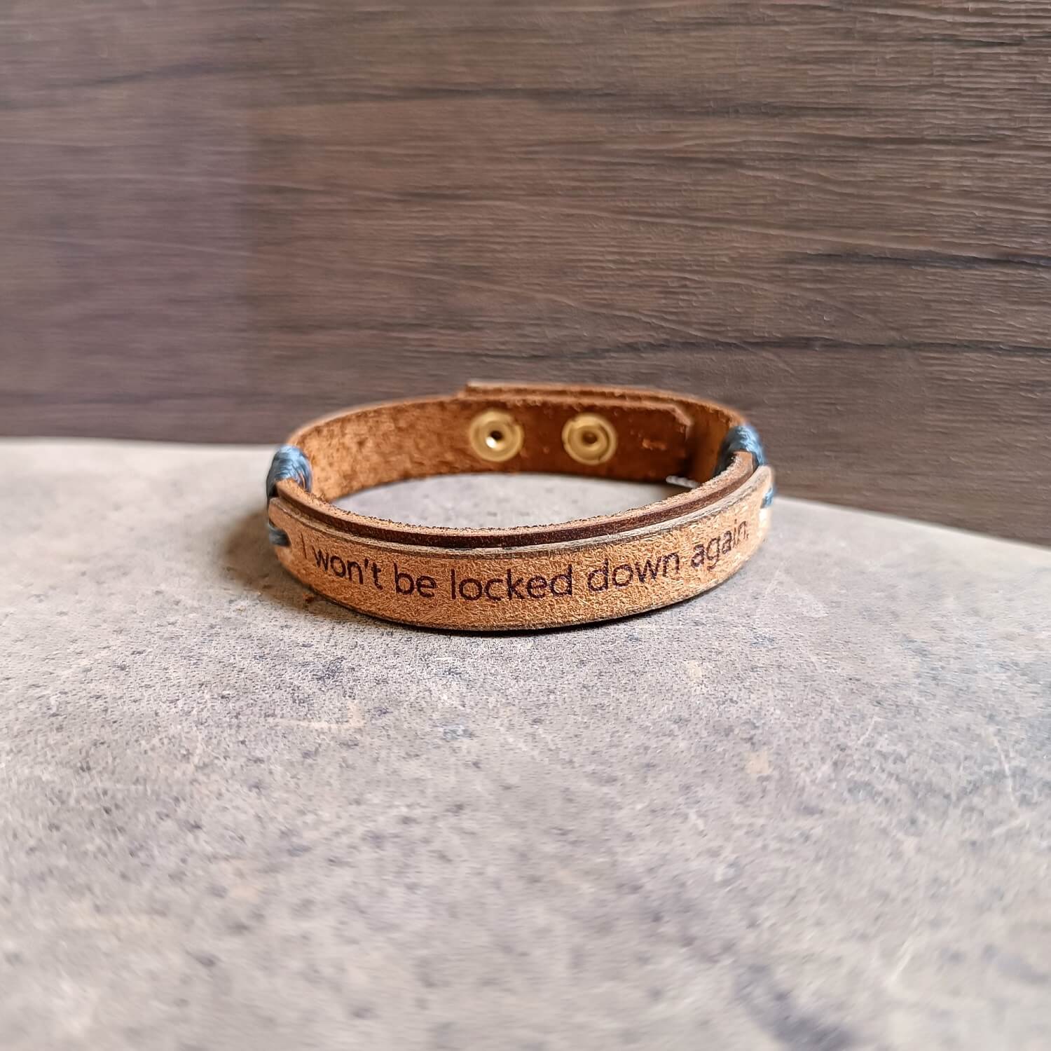Dark brown and beige leather cuff engraved with “I won’t be locked down again,” adjustable stainless steel buttons, close-up side view showing texture, layers and cord