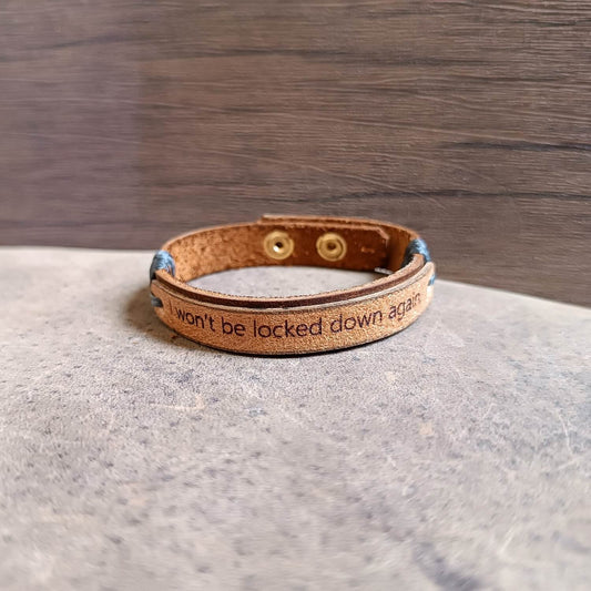 Dark brown and beige leather cuff engraved with “I won’t be locked down again,” adjustable stainless steel buttons, close-up side view showing texture, layers and cord