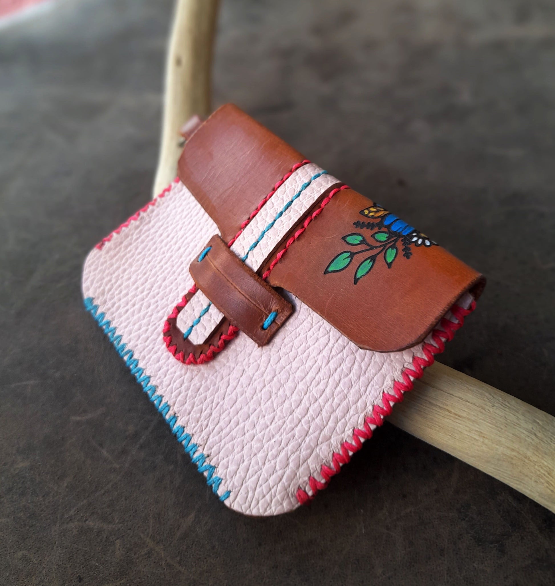 A powder pink leather flap wallet with a brown flap and blue floral hand-painted design, resting on a wooden surface.