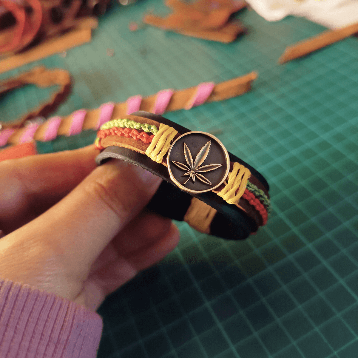 Hand holding a bracelet with a cannabis leaf design on a green cutting mat.