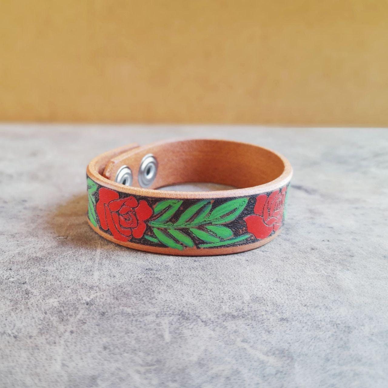 A brown leather cuff bracelet with hand painted red rose vine design, displayed on a textured background.