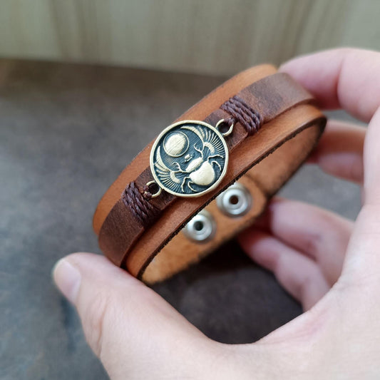 Brown leather bracelet with a winged scarab pendant held in a hand.