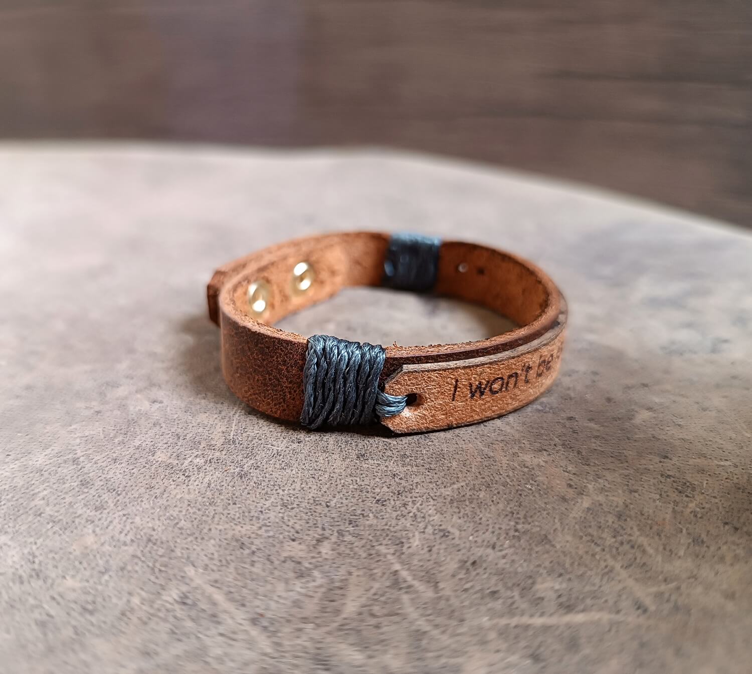 Dark brown and beige leather cuff engraved with “I won’t be locked down again,” adjustable stainless steel buttons, close-up side view showing texture, layers and gray cord