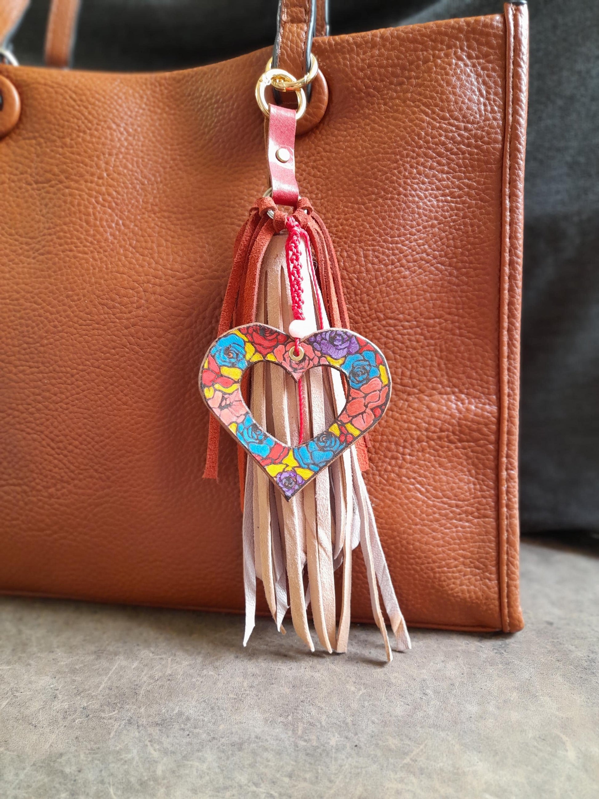 Hand painted floral leather heart-shaped bag charm with a pink leather tassel and red fringe, attached to a brown leather tote bag.
