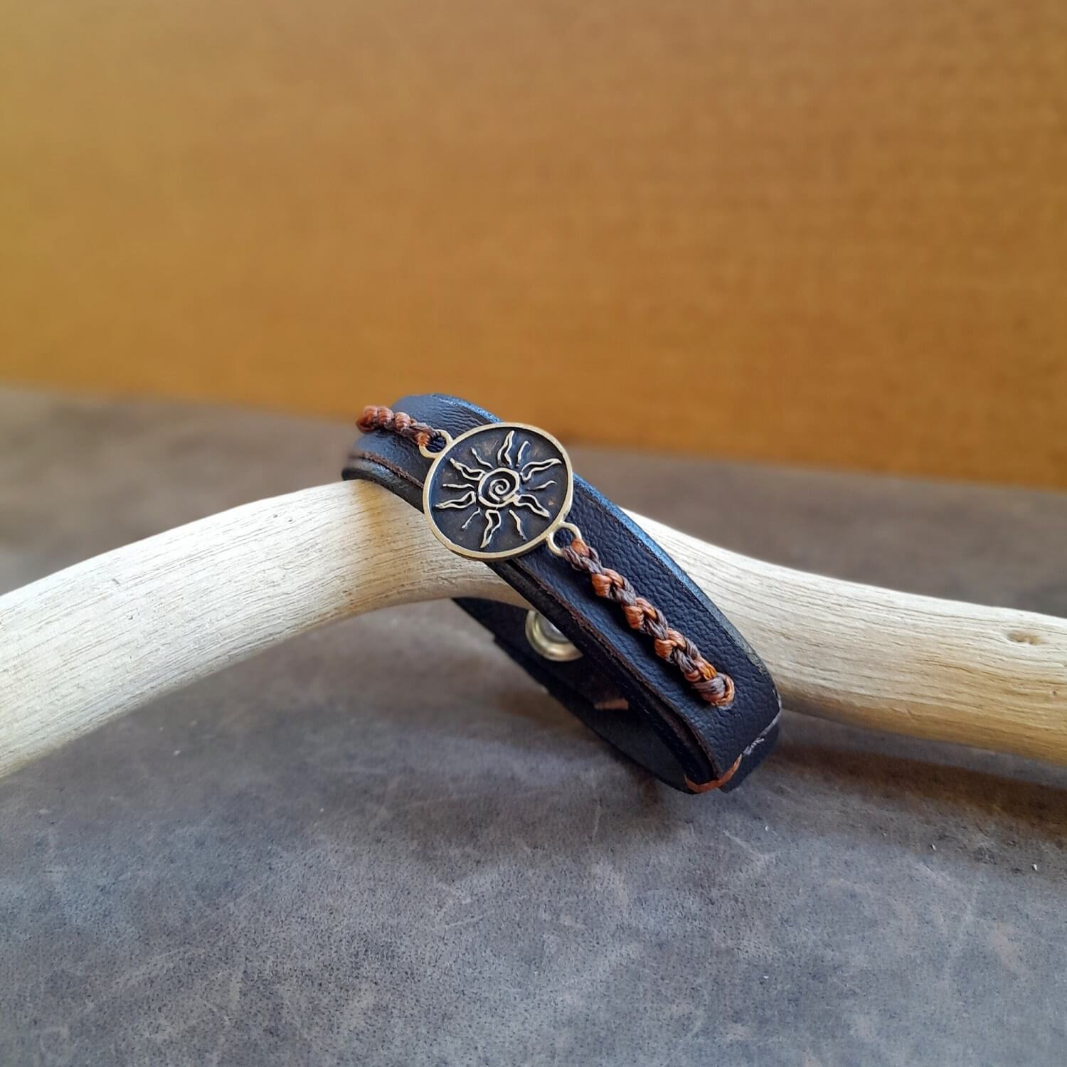 A brown leather bracelet with a handcrafted antique brass Spiral Sun Pendant, decorated with earthy macrame, displayed on a pale background.