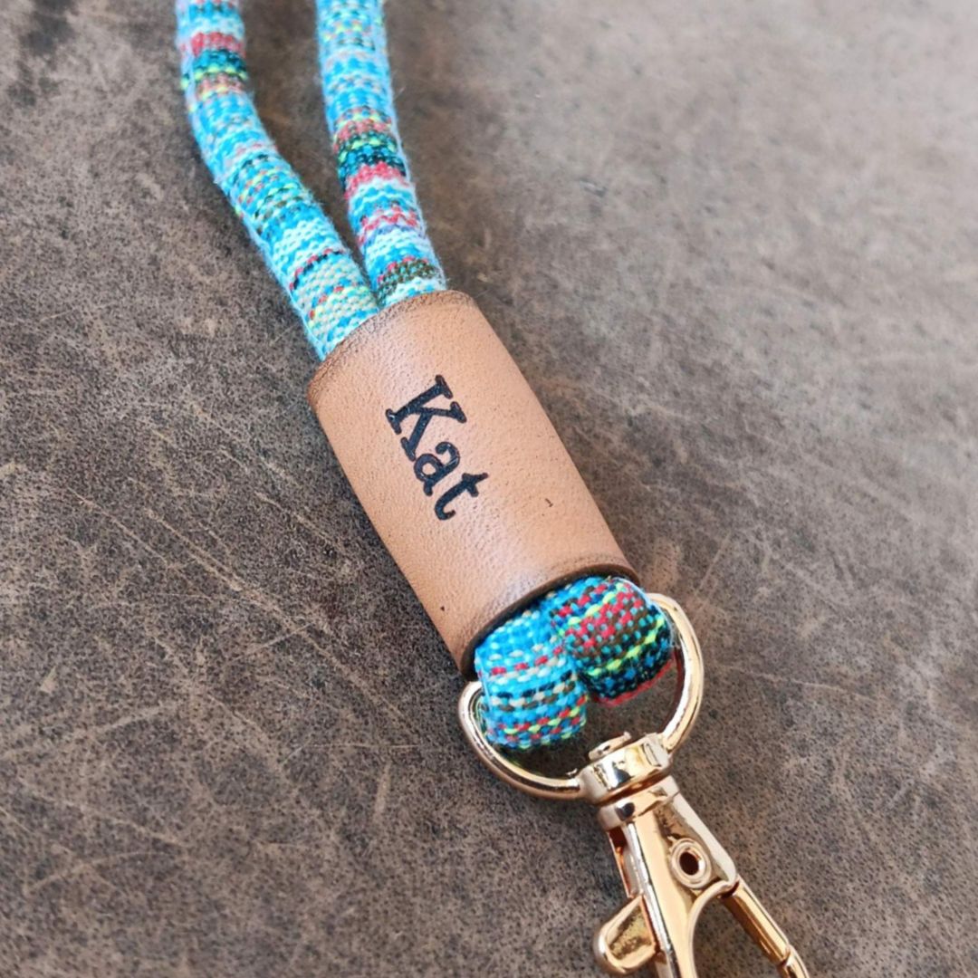A close-up of a personalized keychain or bag strap with a woven blue, red, and green cord, a tan leather wrap engraved with the name 'Kat', and a gold-tone metal clasp, set against a textured dark gray background.
