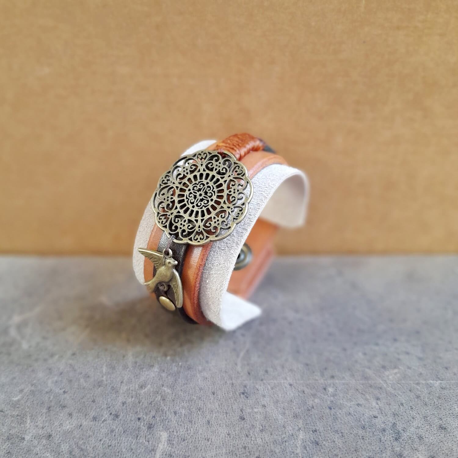 The OOAK "Happy Beige" Mandala Cuff, other side view: a wide leather and suede bracelet in neutral, earthy tones, featuring a large antique bronze mandala flower motif and a small bird charm.
