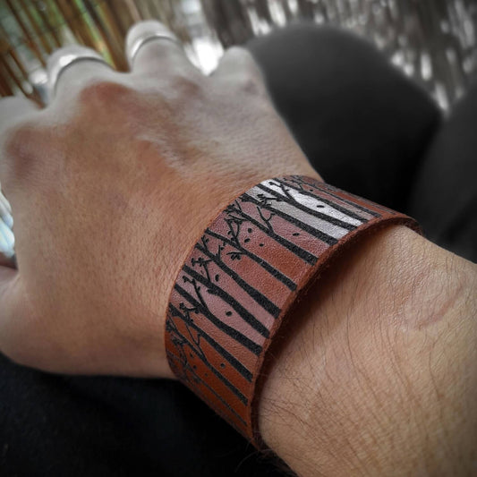 Engraved leather cuff with tree silhouettes worn on wrist, styled with dark outfit.